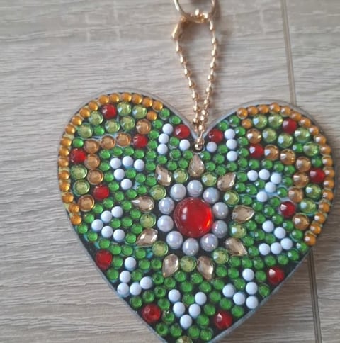 Heart-shaped ornament decorated with multicolored rhinestones and beads in green, red, white, and gold, featuring a large red crystal center, with a rose gold chain attached