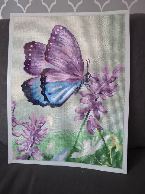 Cross-stitch artwork of a purple and blue butterfly resting on pink flowers with green stems on a cream background