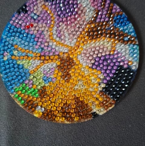 Circular beaded craft with colorful rhinestones arranged in a radial pattern featuring blue, purple, gold, green, and other vibrant hues on a gray surface