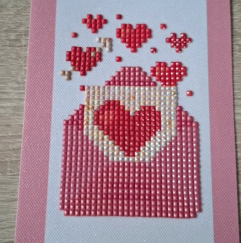 Cross-stitch embroidery of a red and white jar with red hearts floating above it on white fabric with pink borders.