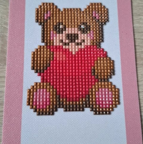 Pixel art teddy bear with brown fur and red shirt on white and pink fabric background