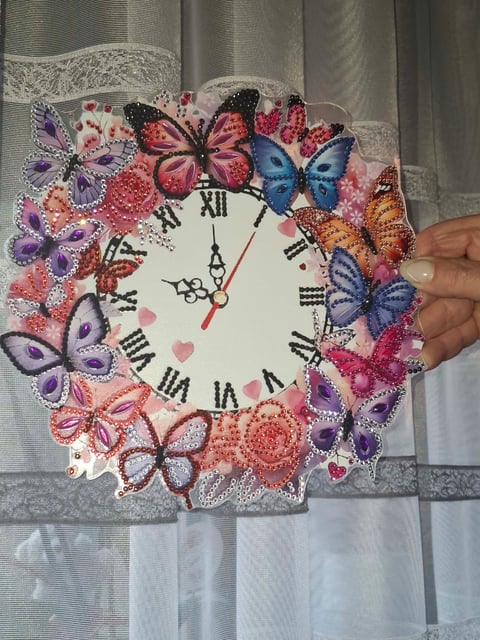 Decorative wall clock with colorful beaded and sequined butterfly border, Roman numerals, held by a persons hand against a lace curtain background