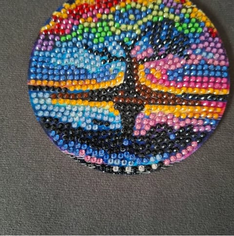 Circular beaded art piece with colorful rainbow segments and black cross pattern in center on gray background