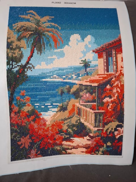 Cross stitch pattern depicting a tropical beach scene with palm trees, blue ocean, sandy shore, red flowers, and coastal buildings