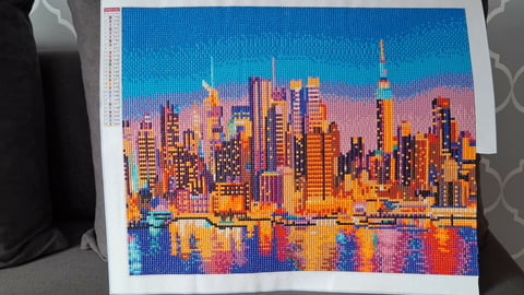 Colorful cross-stitch artwork depicting a vibrant city skyline with illuminated skyscrapers reflected in water below