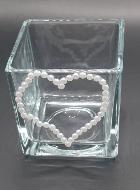 Clear glass cube vase containing a heart-shaped pearl beaded bracelet and pearl jewelry on gray background