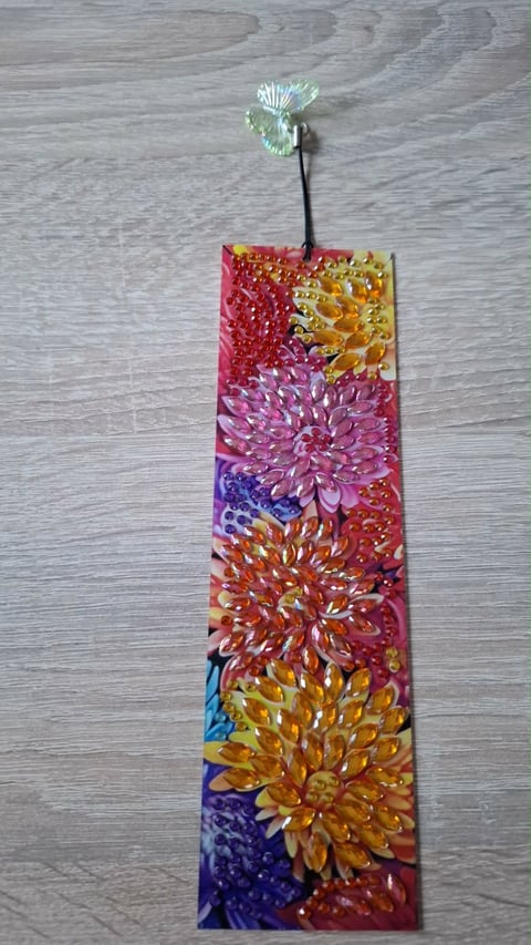 Colorful bookmark with printed dahlia flowers in red, pink, purple, yellow, and orange, displayed with a glass flower charm on wooden surface