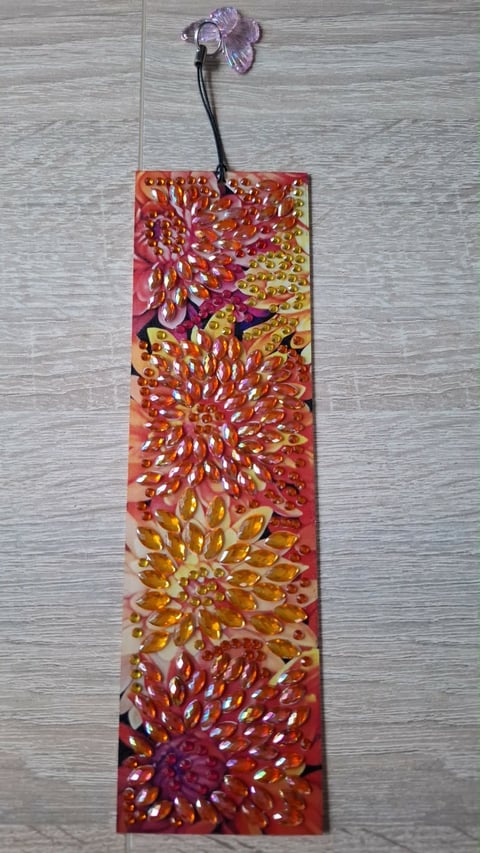Vertical decorative bookmark with colorful painted flower designs in red, orange, and yellow, attached with a purple flower-shaped charm to a wooden textured background