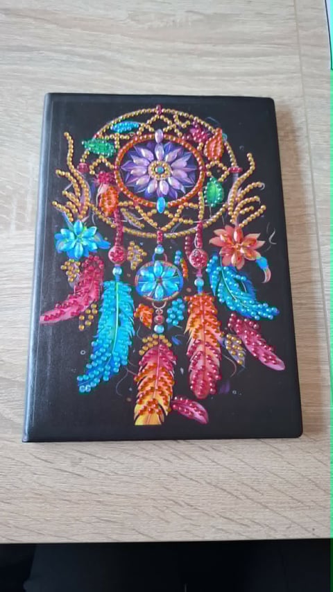 Colorful rhinestone dreamcatcher art on black notebook cover with purple flower, beaded feathers, and hanging ornaments